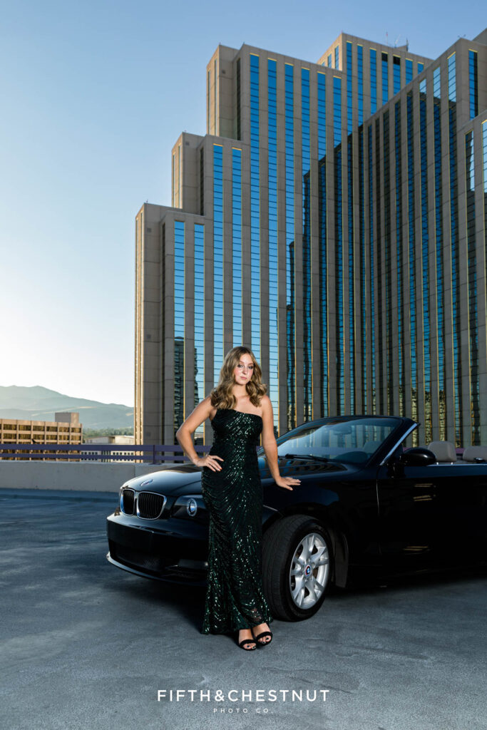 Edgy and modern senior portraits of a female senior wearing an elegant and sparkly prom dress set against the backdrop of a towering Hotel Casino at Golden Hour next to a black BMW convertible.