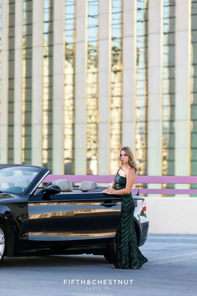 Edgy and modern senior portraits of a female senior wearing an elegant and sparkly prom dress set against the backdrop of a towering Hotel Casino at Golden Hour next to a black BMW convertible.