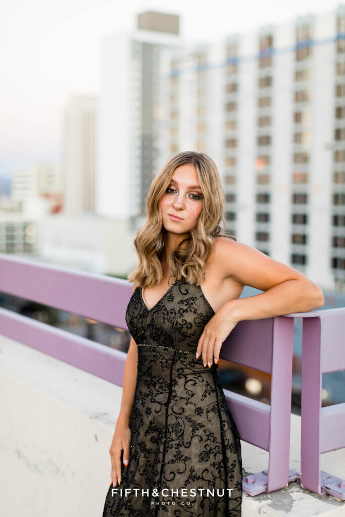 A senior in a black prom dress leans against the parking garage wall overlooking a Downtown Reno Casino for her edgy and modern senior portraits at golden hour with Reno Senior Photographer Fifth and Chestnut.