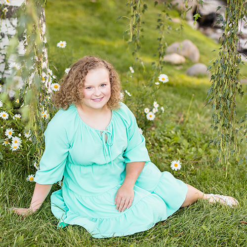 A lovely Bishop Manogue High School Senior in a turquoise flowing dress and stylish sandals sits on grass amidst willow tree leaves and white daisies for a stunning senior portrait by Reno Senior Photographer Fifth and Chestnut Photo Co.