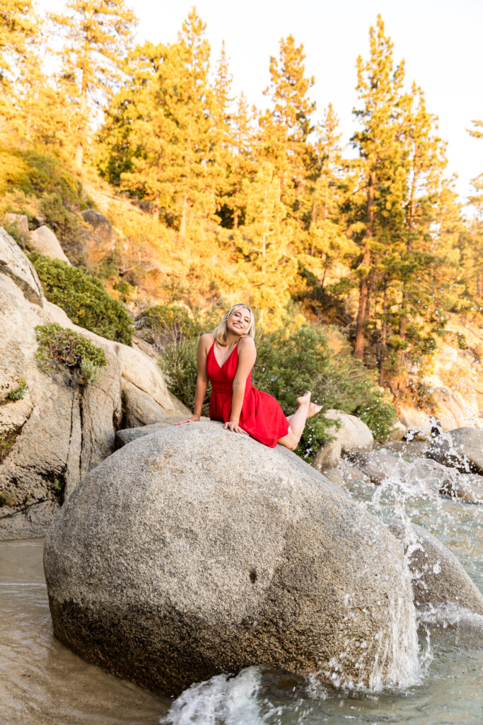 Little Mermaid vibes for this high school senior who is laying on a rock in a red sundress with water splashing up from behind her for her summer Lake Tahoe senior portraits.