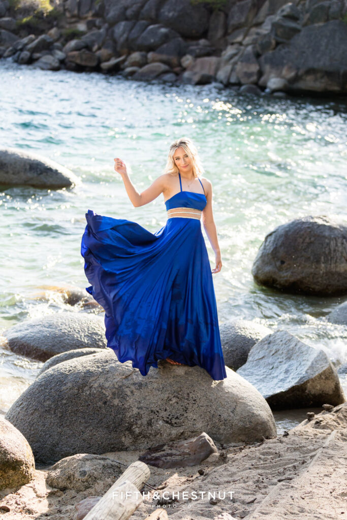 A high school senior girl with blonde hair plays with her lovely blue satin dress as she stands on the huge boulders of the Lake Tahoe shoreline at Secret Cove for her elegant summer senior portraits at Lake Tahoe by Lake Tahoe Senior Photographer Fifth and Chestnut Photo Co.