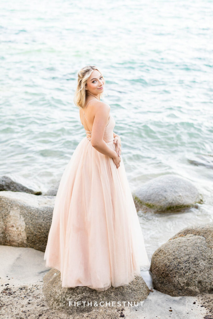 Girl in a blush gown stands on a rock against the backdrop of Lake Tahoe for her elegant summer senior portraits at Lake Tahoe by Lake Tahoe Senior Photographer.