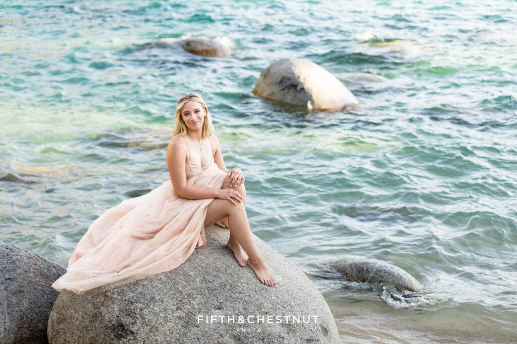 A senior girl wearing a pink sparkly gown sits on a boulder with the blue and emerald water of Lake Tahoe shining behind her for elegant summer senior portraits at Lake Tahoe by Lake Tahoe Senior Photographer