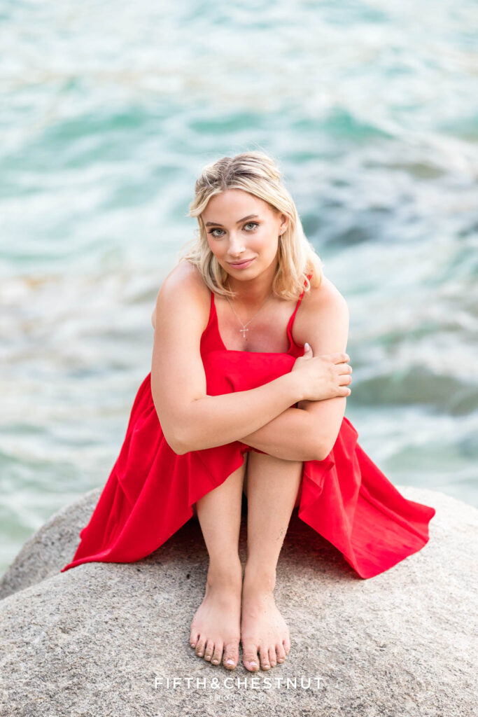 Stunning Lake Tahoe senior portrait of a blonde female wearing a bright red sundress with the blue hues of Lake Tahoe behind her.