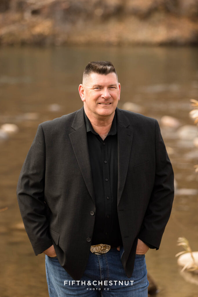 Classic fall outdoor headshots by Reno Headshot Photographer at Mayberry Park of man wearing a black jacket, a black button-up, jeans and cowboy boots by the river.
