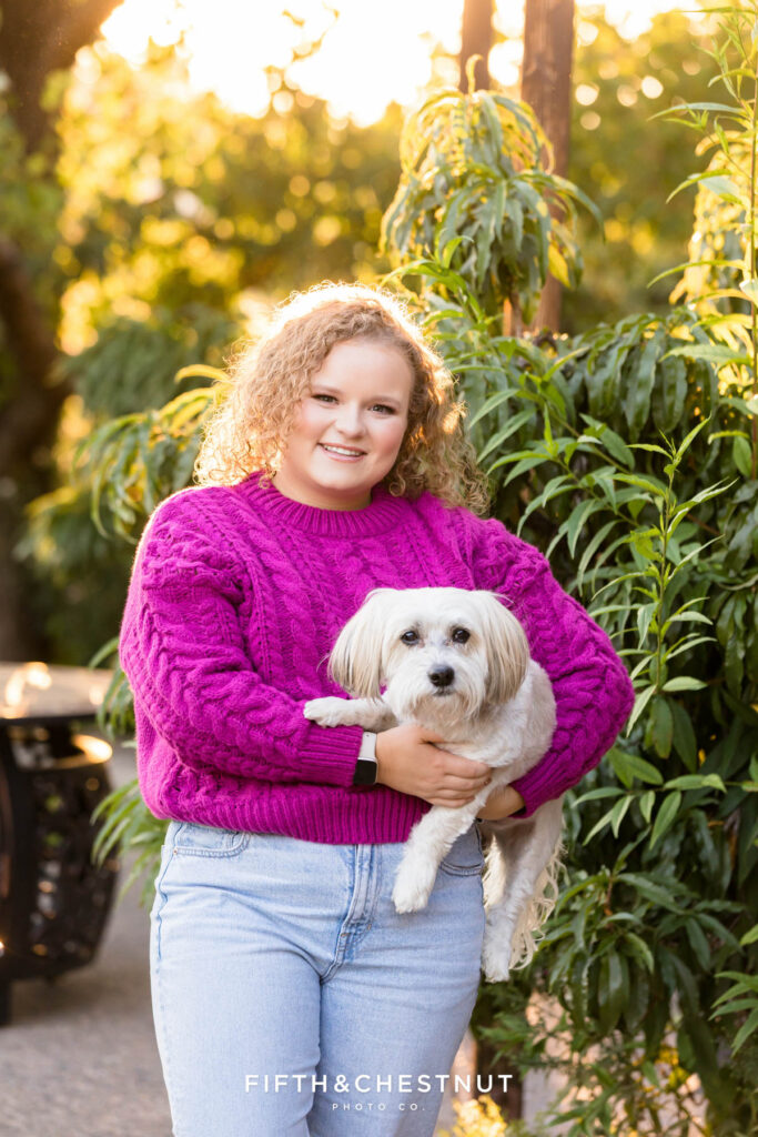 Stunning senior portrait of a Manogue High School Senior by a Reno Senior Photographer holding her sweet puppy with green foliage in the background and the sun shining from behind them both
