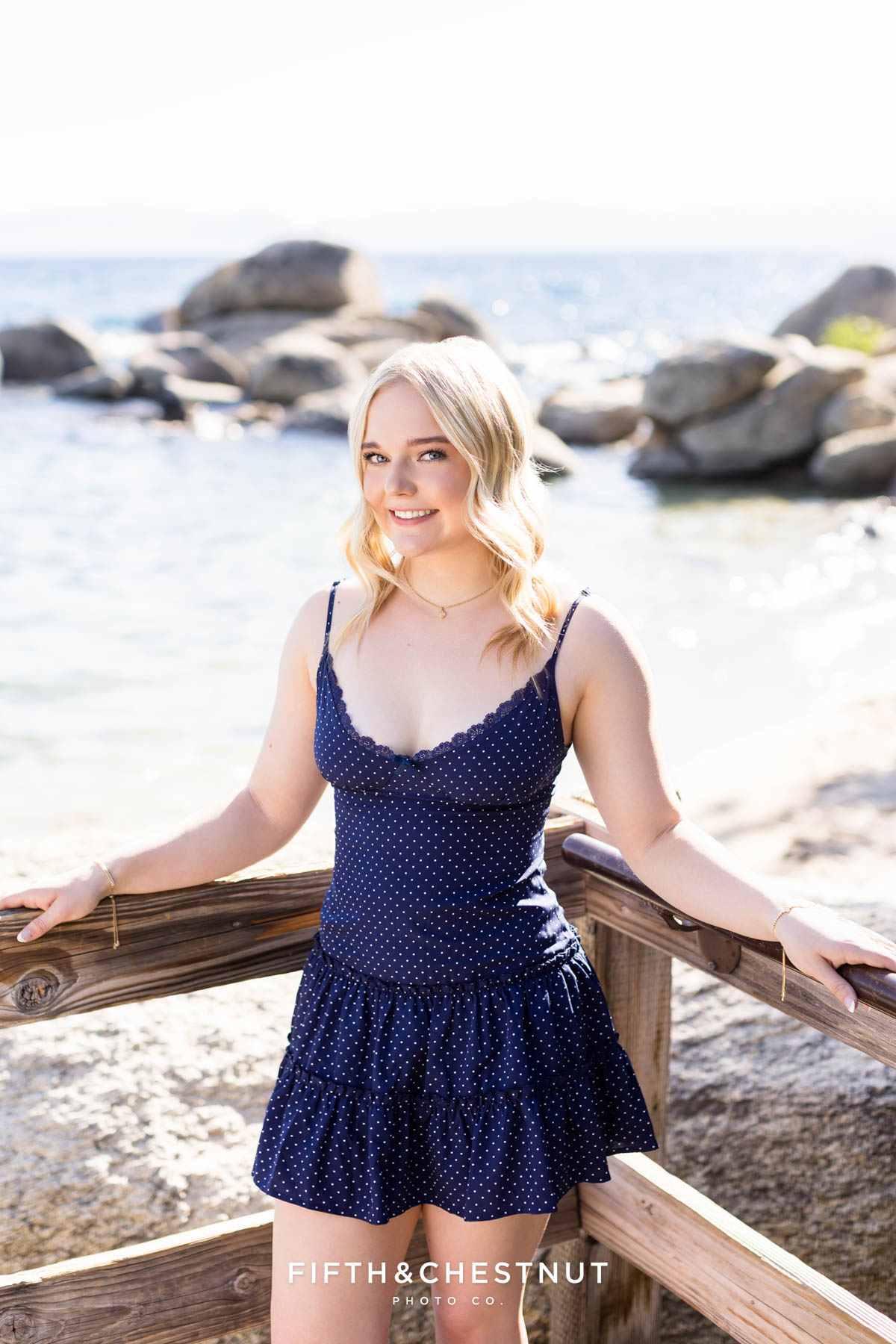Bishop Manogue Senior smiling by a rustic wooden fence surrounded by bright shimmery lake water behind her.