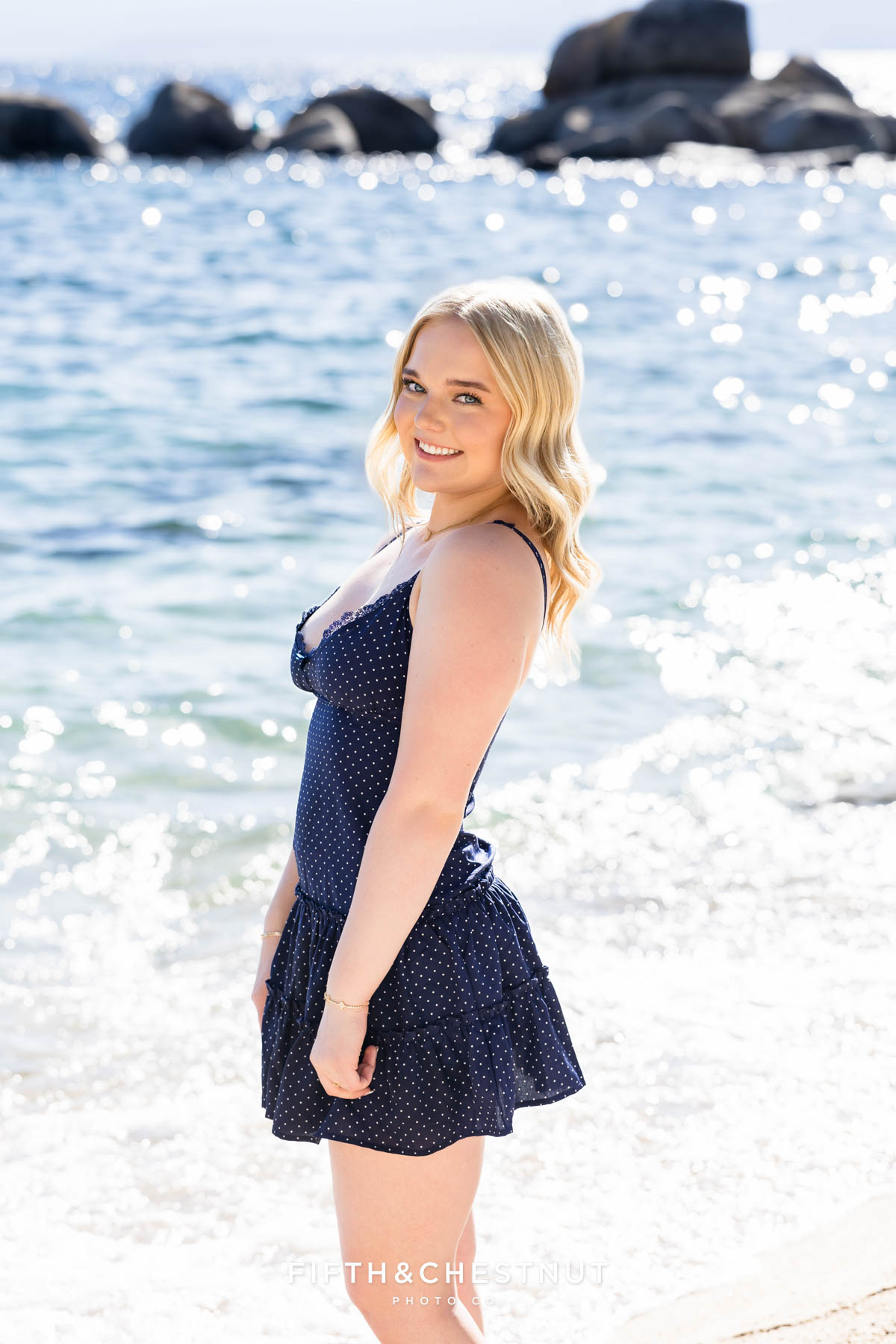 Bishop Manogue Senior standing in front of an outcrop of boulders on lake tahoe with the shimmery blue water sparkling all around her