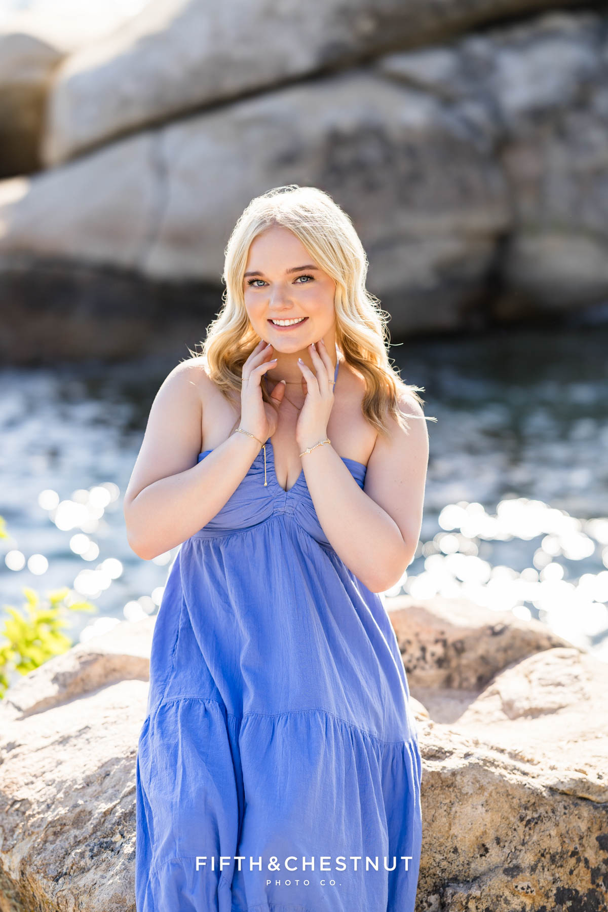 Bishop Manogue Senior in a flowing blue dress sitting on large granite boulders in Tahoe with the sparkling lake below.
